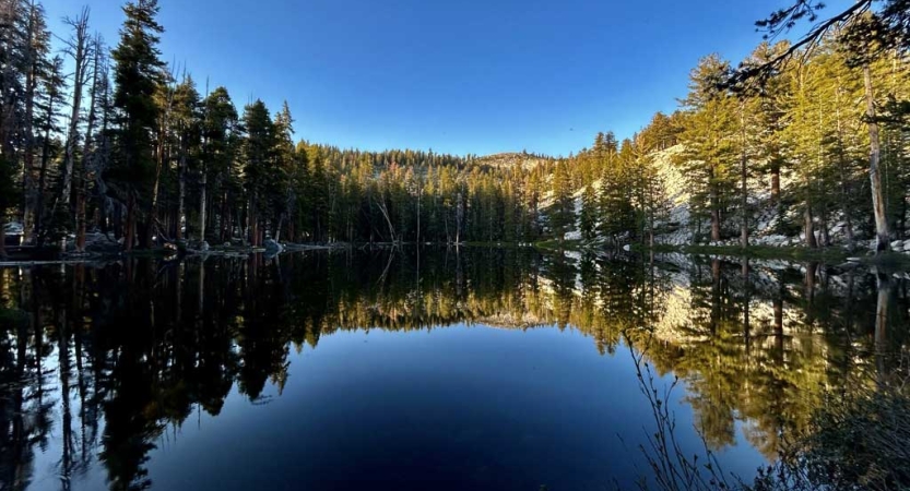 A lake reflects the evergreen trees, hills and blue sky that frame it. 